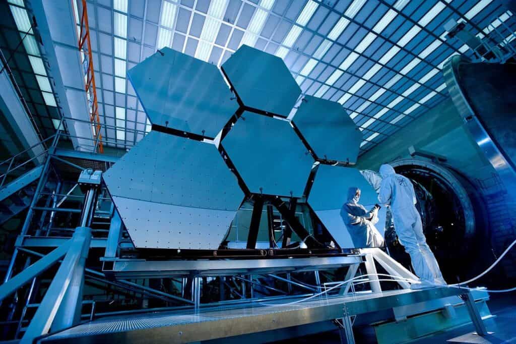 Two technicians in cleanroom suits, under the blue laboratory lighting, meticulously examine a large hexagonal mirror array. Their precision reflects the dedication akin to Rockwell Automation's standards.