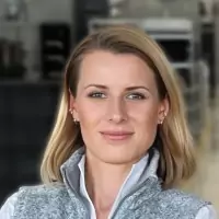 A woman with blonde hair is facing the camera, wearing a gray zip-up vest over a white top. The background appears to be an indoor, modern environment with blurred details.