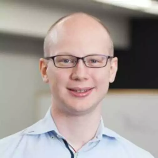 A person with short hair, glasses, and a light blue shirt is smiling at the camera in an indoor setting.