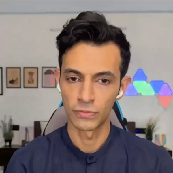 A person with dark hair wearing a dark shirt and wireless earbuds sits indoors with a neutral expression. The background includes wall art and colorful triangular lights.