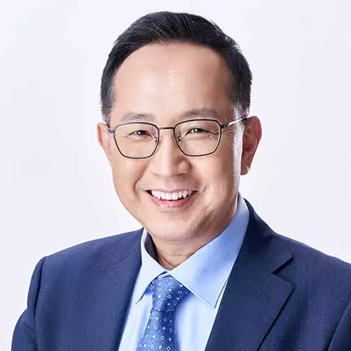 A man wearing glasses and a blue suit with a dotted tie smiles at the camera against a plain background.