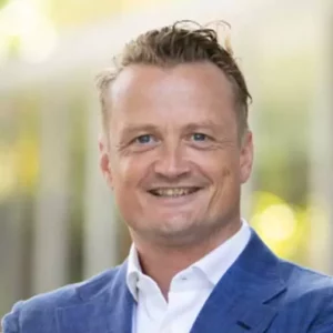 A man with short hair in a blue suit and white shirt smiles in an outdoor setting with blurred greenery in the background.