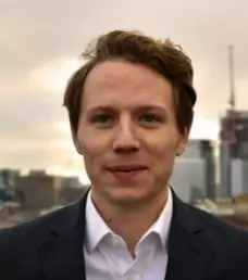 A person with short hair and a slight smile is shown outdoors with a cityscape in the background. They are wearing a dark jacket and white shirt.