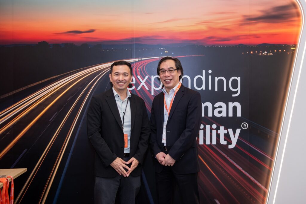 Two men smiling in a conference room, standing in front of a banner with the text "expanding human possibility," with a backdrop of a highway at sunset.