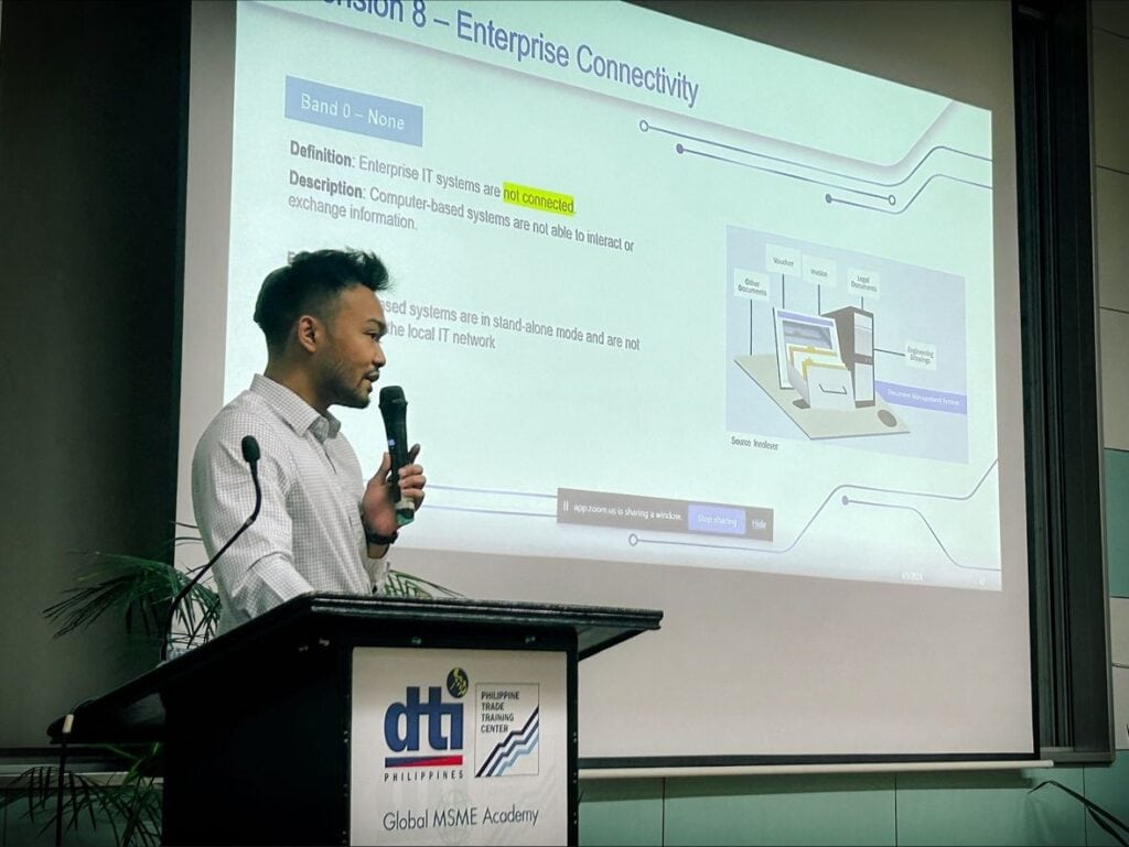 A man in a white shirt is giving a presentation at a podium with a microphone. A slide on Enterprise Connectivity and SIRI training is displayed on the screen behind him.
