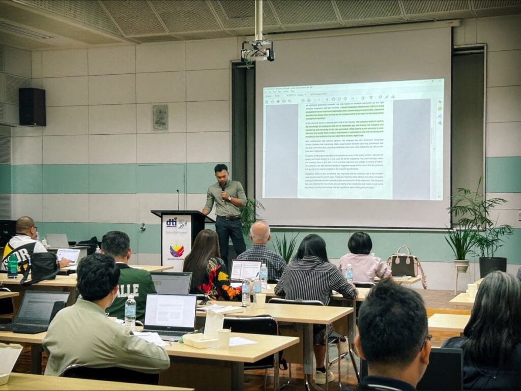 A man stands at a podium in a classroom, presenting a document projected on a screen. Several people are seated at tables with laptops, intently following the SIRI training session.