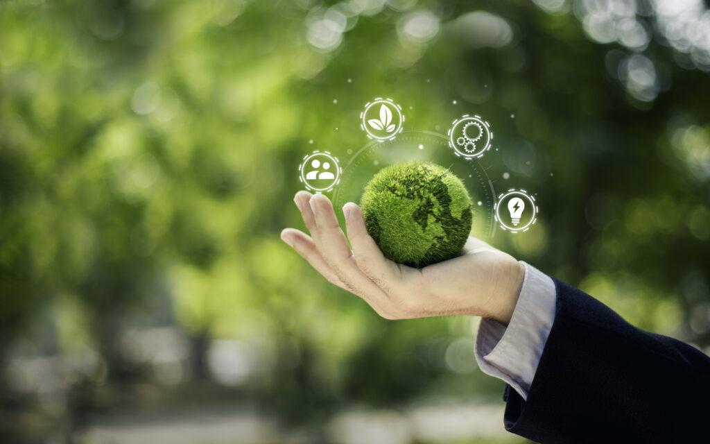 A hand in a suit holds a green mossy sphere with environmental icons floating above it, set against a blurred natural background, symbolizing the harmony between manufacturing and nature.
