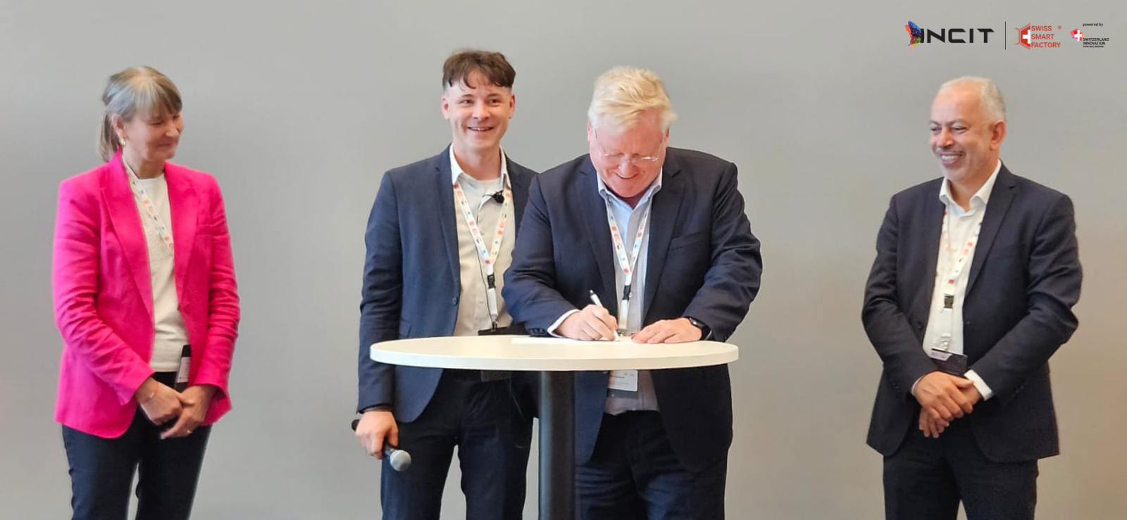 Four people stand around a round table, one man is signing a document while the others watch and smile. Logos are displayed in the upper right corner.