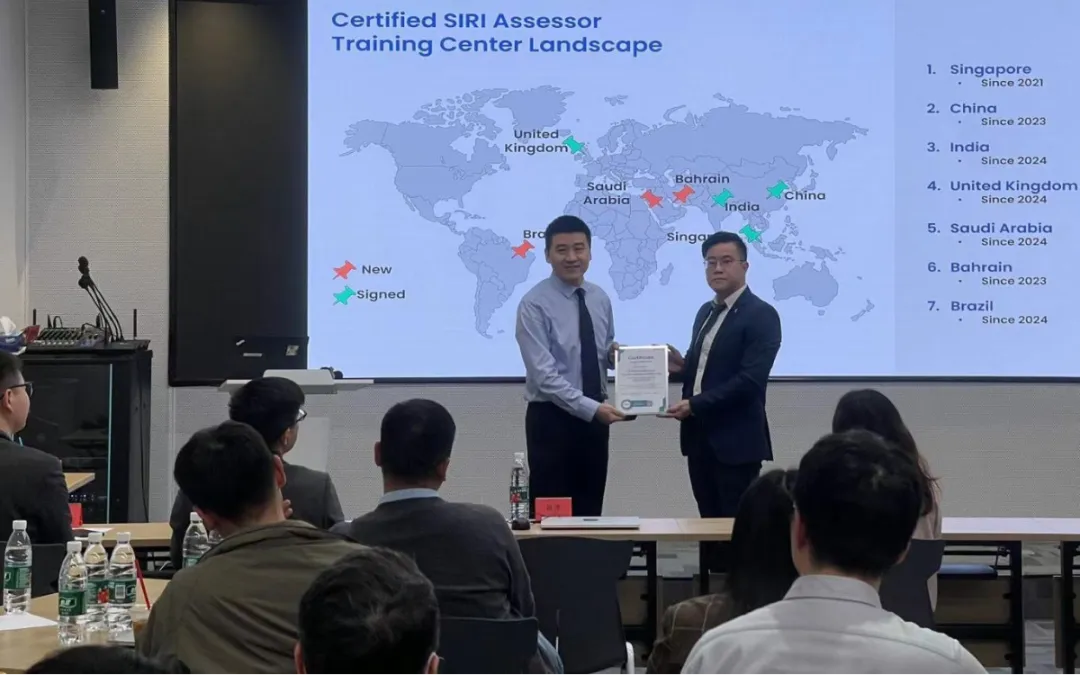 Two men are standing at the front of a classroom, facing the audience. One man is handing a certificate to the other. A presentation slide in the background maps countries with certified assessors.