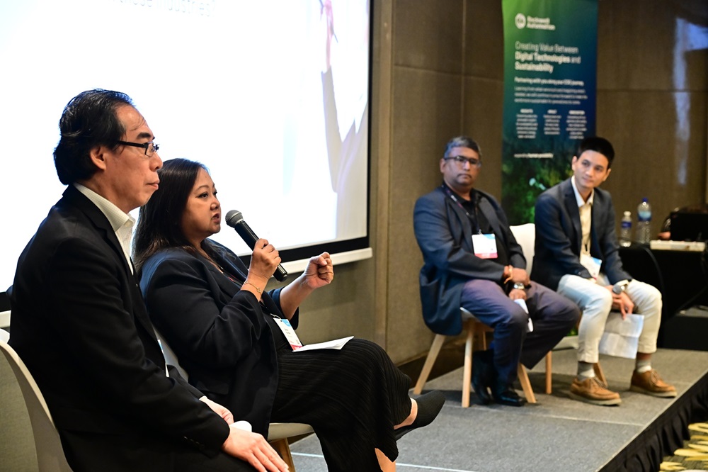 Four individuals sit on a panel in a conference room; one woman speaks into a microphone while the others listen attentively. A presentation slide showcasing Rockwell Automation is partially visible in the background.