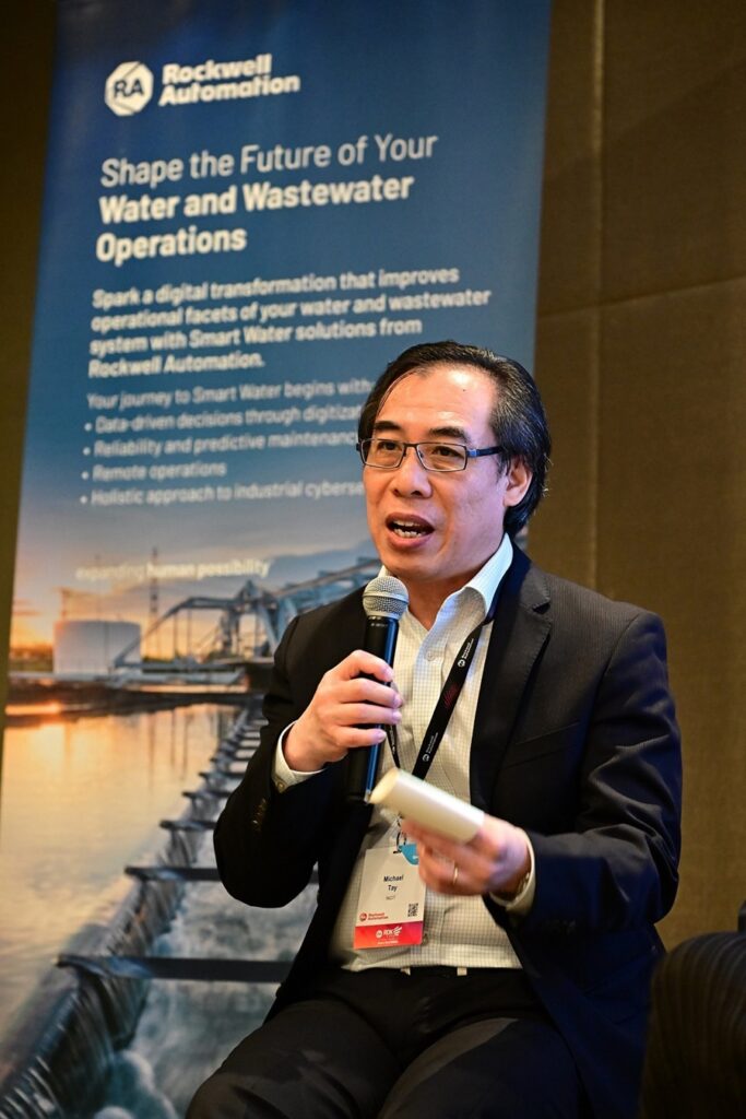 A man in a suit speaks into a microphone while holding notes. Behind him, a banner reads "Shape the Future of Your Water and Wastewater Operations" with details about Rockwell Automation solutions, highlighting their innovative approach to managing essential services efficiently.