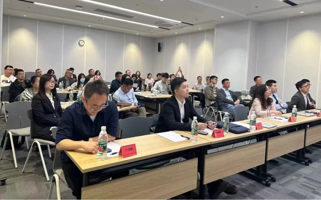 People sitting at desks in a conference room, listening to a speaker. Some are taking notes, and water bottles are visible on the desks.