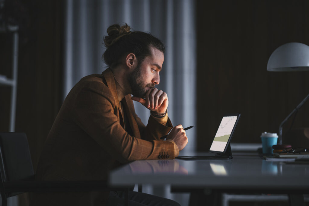 A man with a bun and beard sits at a desk, focused on a tablet screen, holding a stylus. The setting is dimly lit, with a lamp in the background and a coffee cup to his right.