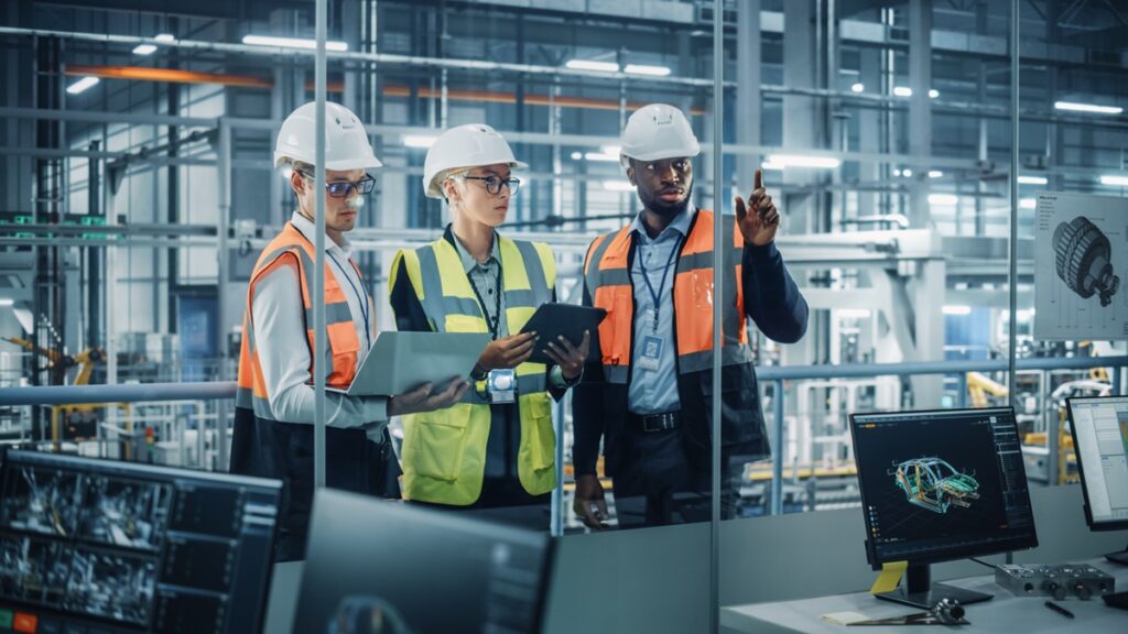 Three engineers in safety gear and hard hats look at data on screens and discuss in an industrial control room. One points at the screen while the others hold laptops. Factory machinery is visible.