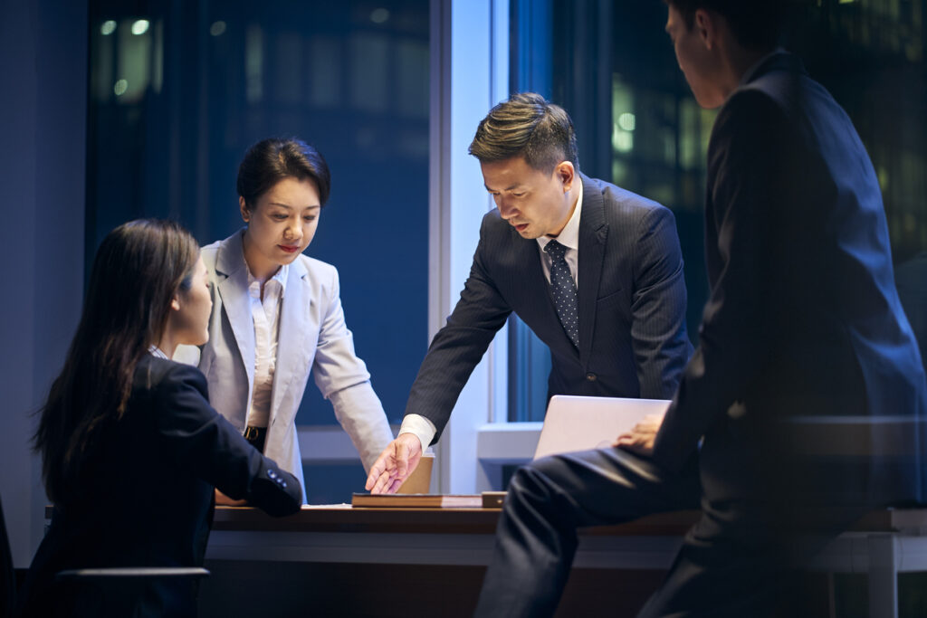 Four people in formal business attire engage in a serious discussion around a desk in an office setting during the evening, focusing on leadership training.