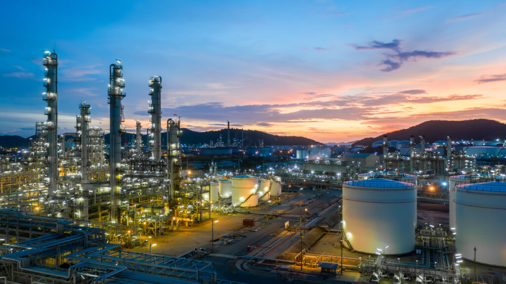 An expansive oil refinery complex with large storage tanks and towering structures is illuminated during twilight, set against a backdrop of hills and a colorful sunset sky.