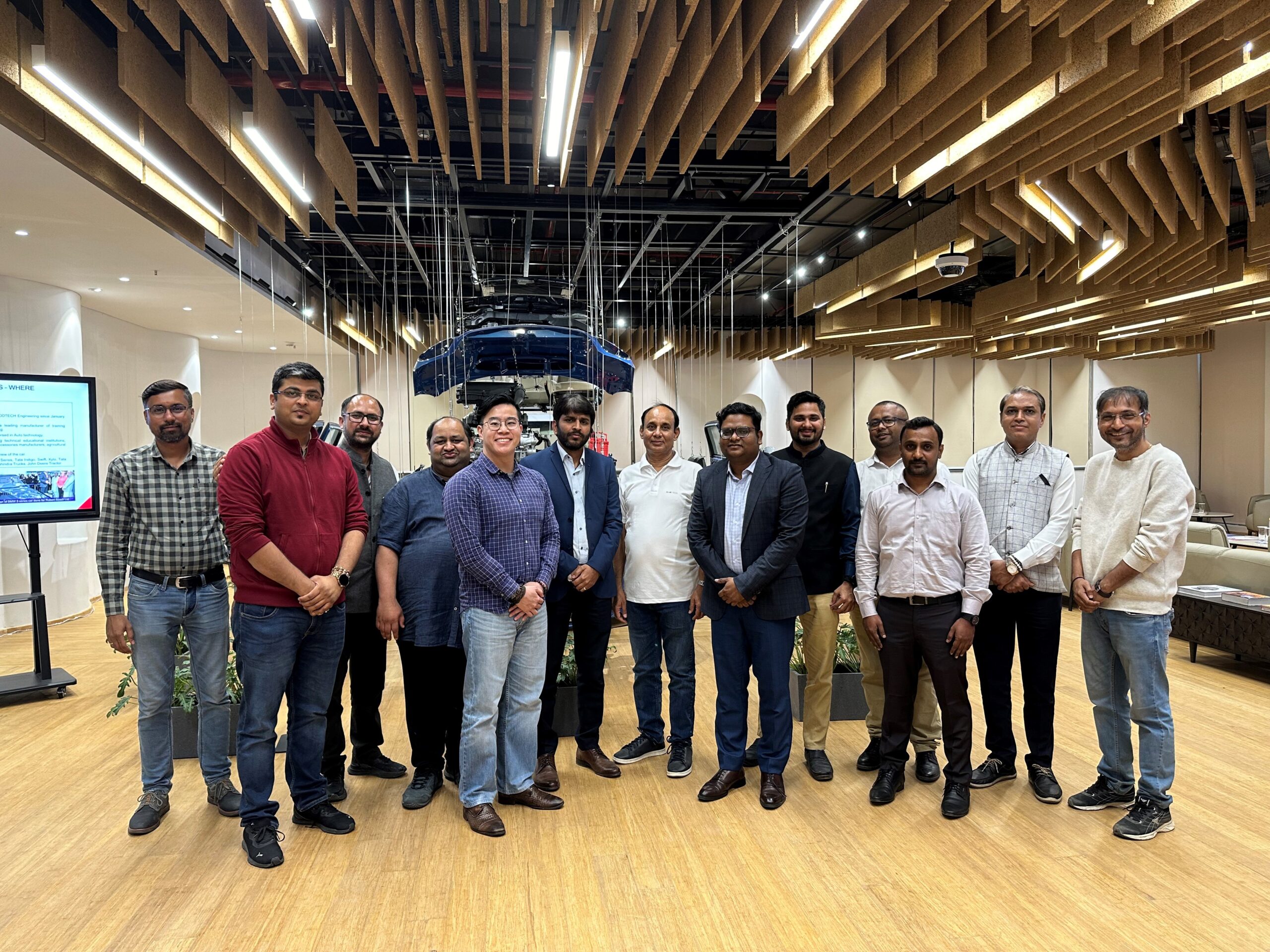 A group of fifteen individuals, mostly men, standing in an office space with a modern ceiling and wood floor. One person is holding a presentation remote. A display screen is visible in the background.