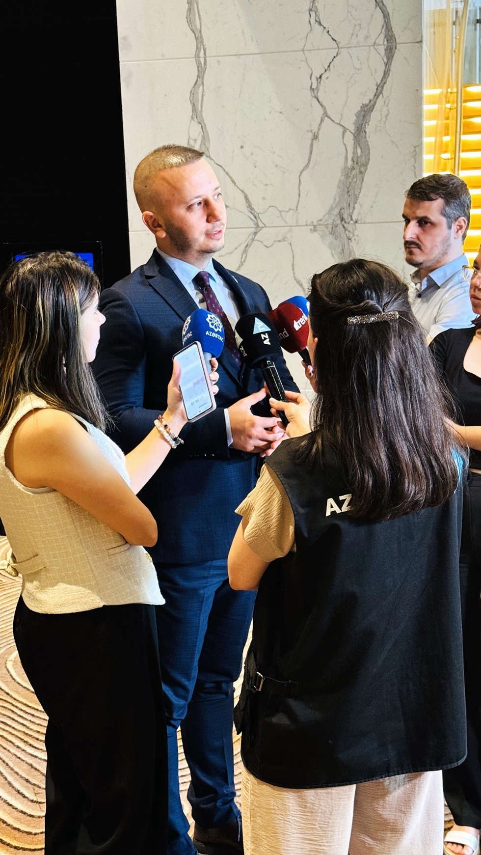 A man in a suit is being interviewed by several reporters, with one holding a microphone and another recording on a smartphone. The setting appears to be indoors with a marble wall in the background.