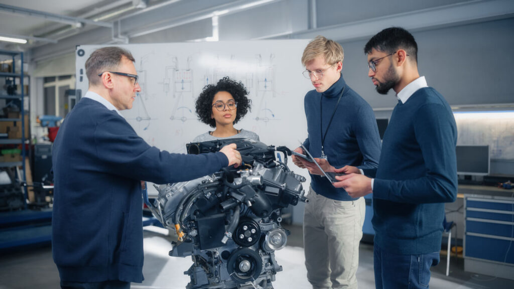 Four people in a workshop focus on an engine model. One person gestures while explaining, and the others observe and take notes. A whiteboard with diagrams is in the background.