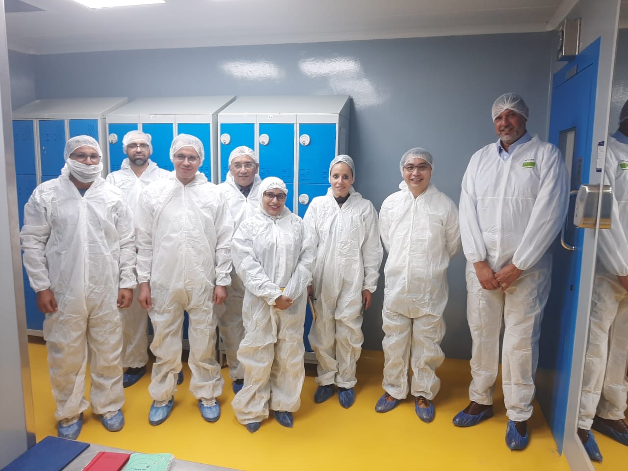 A group of people wearing white protective suits, blue shoe covers, and hairnets standing in a cleanroom environment with lockers in the background.