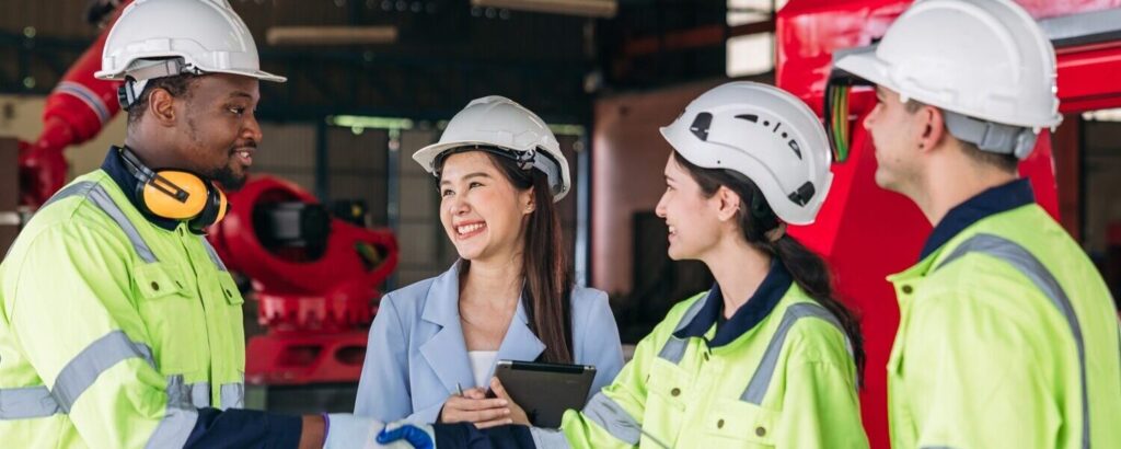 Four people in safety gear and business attire stand together in an industrial setting, shaking hands and smiling. Two individuals hold tablets, likely discussing strategies for mitigating physical risks.