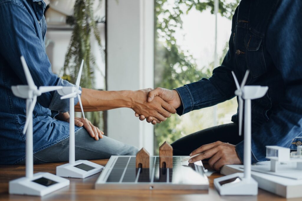 Two people shaking hands over a table with model wind turbines and solar panels, suggesting a discussion or agreement on renewable energy projects.