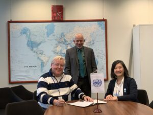 Three people meet at a table with UNIDO flag; a world map is on the wall in the background.