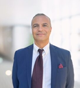 Man in a blue suit and red tie smiling while standing indoors.