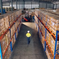 A warehouse worker in a yellow safety vest walks between tall shelves stacked with boxes, while an ethical forklift operator ensures safe and responsible handling in the background.